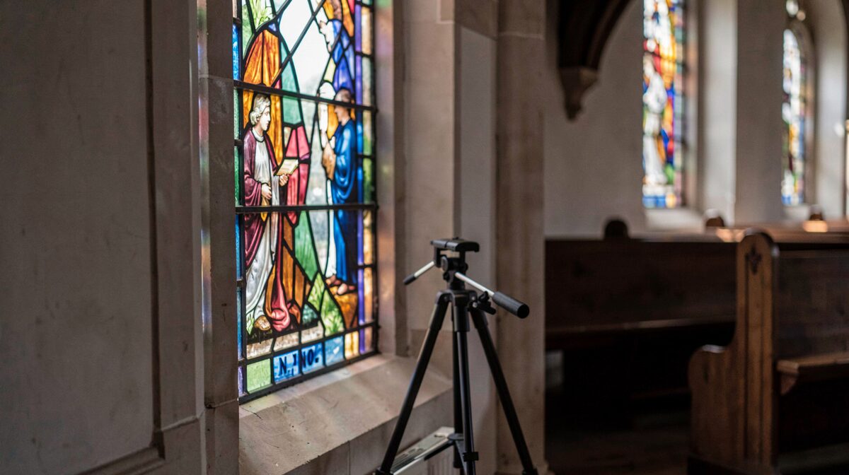 Binnen in een kerk, een glas-in-loodraam van dichtbij gefotografeerd vanaf een statief, zacht daglicht dat door het glas valt, houten banken op de achtergrond in de schaduw, geen flits.